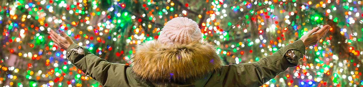This is a picture of a person in a green coat with a flffy hood with arms spread in front of commercial bright Christmas lights.