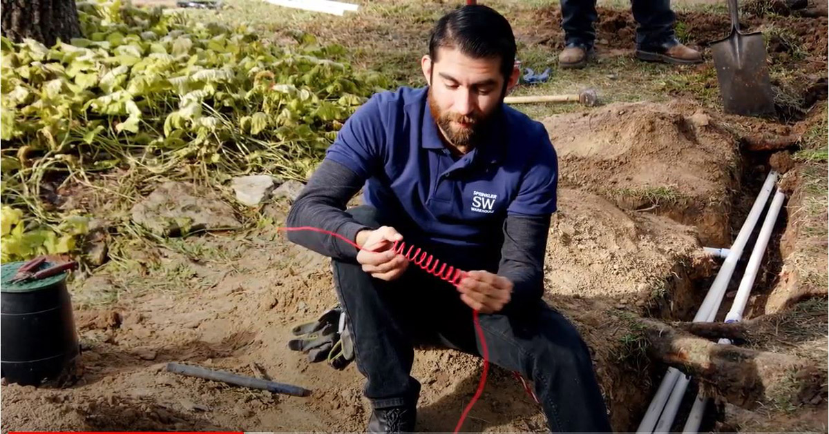A thumbnail of the sprinkler warehouse pro holding a red sprinkler wire while sitting next to a trench with irrigation pipe laid in it.