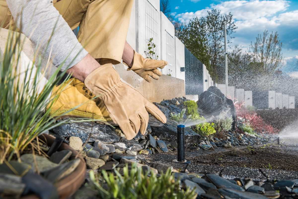 Eco-Friendly Gardens: Grow a Greener Tomorrow with Your Home Ecogarden 3 Man in khaki gloves and pants adjusting a sprinkler rotor irrigating an elaborate garden of rocks and plant life adjacent to a border wall.