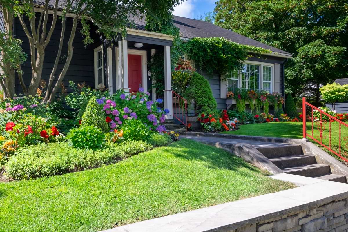Eco-Friendly Gardens: Grow a Greener Tomorrow with Your Home Ecogarden 1 Modest, blue middle class home with a red door and white-trimmed windows behind a sloped landscape festooned with elaborate, colorful gardens and plants in the front yard.