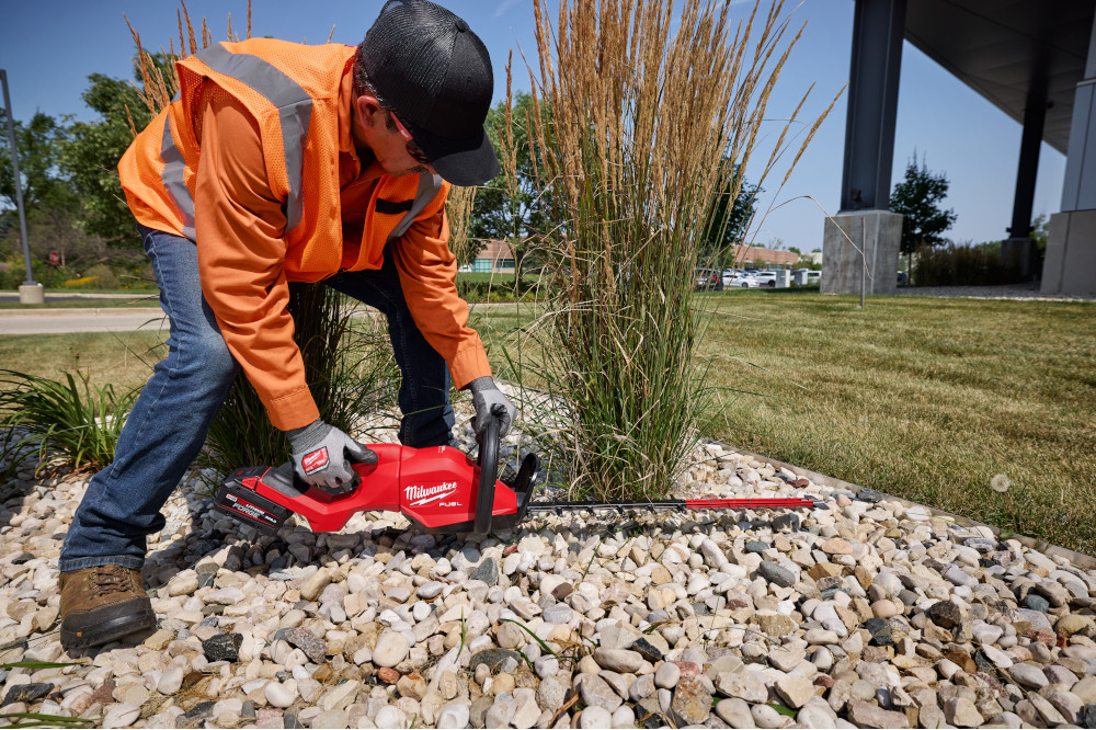 Maintenance 1 Man in orange vest and construction gloves using a Milwaukee trimmer to cut a dogtail plant.