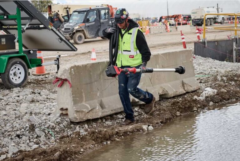 Roadside construction worker walking through mud and water on a jobsite holding a Milwaukee Tool stick transfer pump.