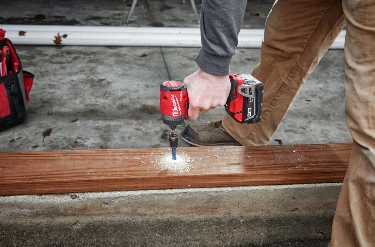 Person bending over drilling a laid-out piece of two-by-four wood using a Milwaukee Tool drill with sawdust collected around the hole as the drill penetrates.