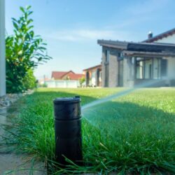 Troubleshooting 2 Sprinkler rotor head spraying water onto a green lawn on the right with a house in the background on a sunny day.